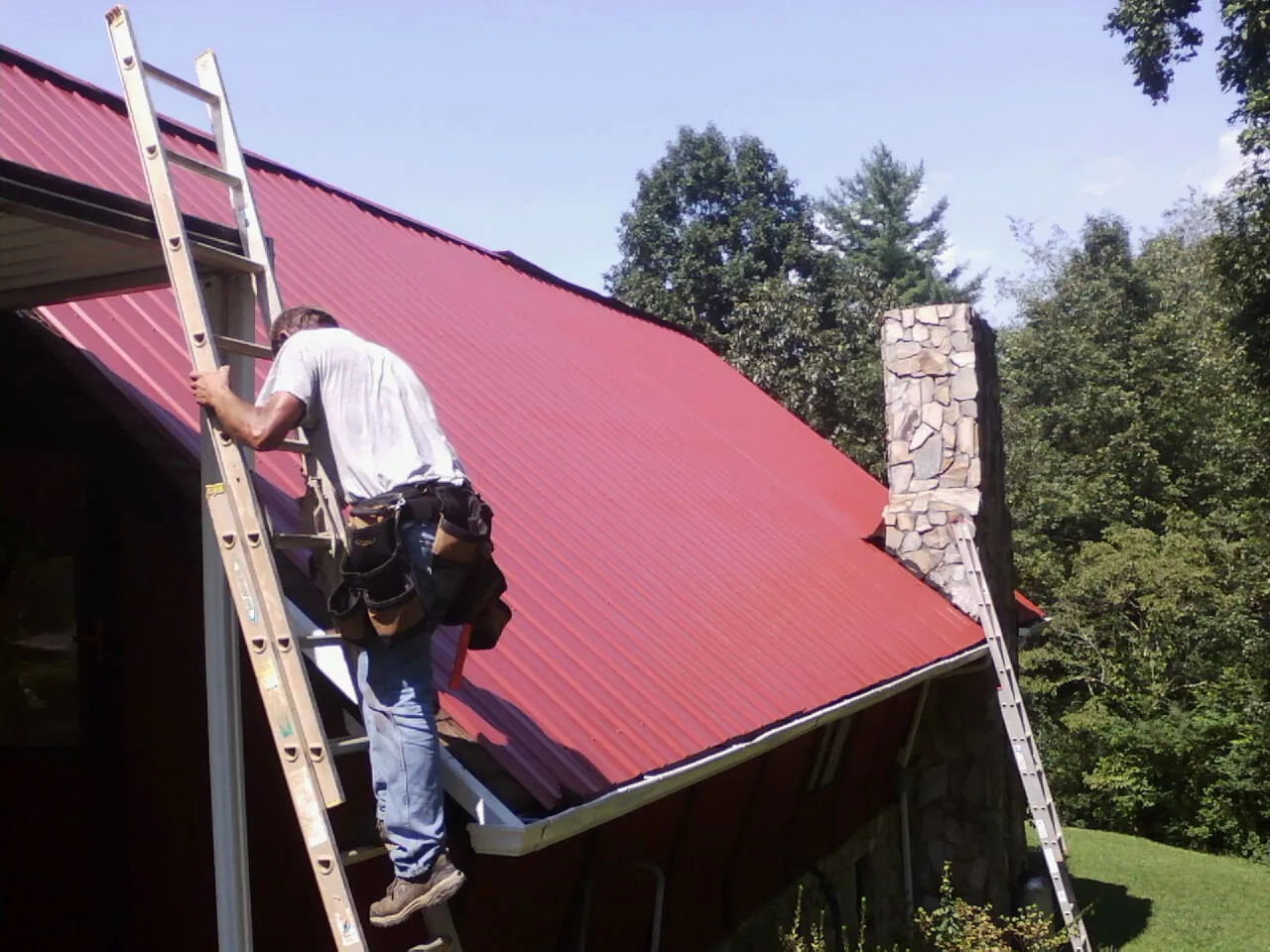 Roofer on a ladder beside a red metal roof and stone chimney — Soffit & Fascia Repair by Roofing Brothers in Silver Lake